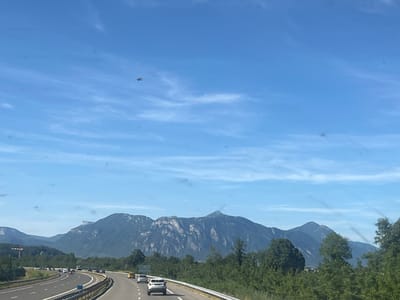 A helicopter is flying in a clear blue sky over a highway in La Buissière, France. The highway has several cars on it. In the background, there are mountains and trees. The scene appears to be captured from a moving vehicle, given the perspective and the slight blur of the landscape. The helicopter is small in the frame, located in the upper left quadrant of the sky.