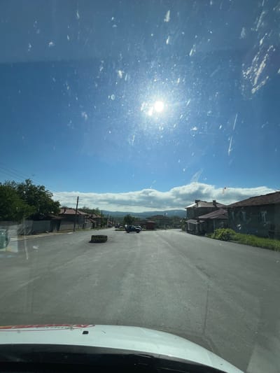 The image depicts a street scene in the city of Rish, Bulgaria, taken from inside a vehicle. The street is paved and asphalt. A discarded tire sits on the right side of the road near the foreground. Buildings line the street, varying in style and age, with trees and foliage visible in front of some of the structures. Overhead power lines run parallel to the street. A vehicle is driving along the street. The sky above is blue with clouds, and the sun glares brightly. The windshield of the vehicle appears to have dirt and streaks. The passenger-side portion of the car is visible to the right side of the image, the red and white car door on the right is of a van.