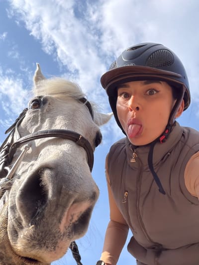 A low-angle, upward-facing close-up shot features a woman wearing a dark helmet and light brown vest, sticking her tongue out at the camera. Beneath her is a white horse with a brown bridle, its head oriented towards the viewer. The background reveals a blue sky with scattered white clouds. The location (Grad Labin, Croatia) is not evident from the image.