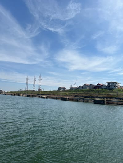 The scene is located in Medgidia, Romania. The foreground consists of a body of dark greenish-blue water, exhibiting surface ripples. A concrete utility structure, featuring yellow conduits, spans horizontally just above the water's surface, separating it from the land. Beyond this structure, a green, grassy embankment slopes upward. On the elevated terrain, a mix of structures is visible. To the left, three metal lattice electricity transmission towers stand prominently. Further left in the distance, industrial buildings are discernible. To the right, a cluster of buildings includes several completed residential-style structures with dark roofs, one building under active construction with exposed concrete framing, and a white-bladed wind turbine. The sky is clear blue with scattered white cirrus clouds. Multiple small, dark avian forms are visible in the upper left sky, and a faint aircraft contrail is present in the upper right. No human figures are depicted.