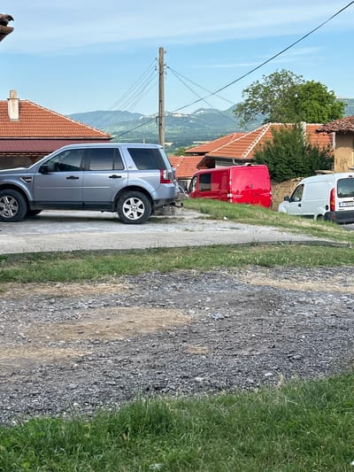 The image depicts a rural scene in Rish, Bulgaria, with several vehicles parked on a gravel and concrete area. In the foreground, a silver SUV is parked on the left. To its right, a bright red van and a white panel van are visible. The background features a village setting with traditional houses having red-tiled roofs, lush green trees, and rolling hills under a bright blue sky with scattered clouds. A utility pole with numerous wires stands prominently in the middle ground. The ground is a mixture of rough gravel and patches of dry grass and soil, with a strip of green grass in the lower part of the frame. The overall impression is a quiet, daytime scene in a Bulgarian village.