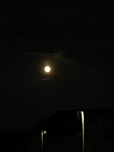 The image depicts a nighttime scene dominated by a dark sky with a bright, full moon. Subtle clouds are visible around the moon, which is located in the upper center of the frame. In the foreground, silhouetted against the night sky, is a building with a dark roofline. A streetlamp stands prominently on the right side of the frame, its light illuminating faint streaks that suggest falling rain or atmospheric disturbance. Windows are visible on the building's facade, some appearing illuminated. The setting appears to be an urban or suburban environment in Wettstetten, Germany, characterized by artificial lighting from the streetlamp and building. No people are visible in the image.