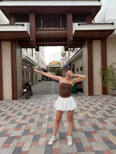 A young woman stands in what appears to be a commercial alleyway in Nghĩa Trụ Commune, Vietnam. She is wearing a brown strapless top and a white bubble skirt, with white sneakers. The woman is posing with her arms extended, her hands forming a V-shape over her face, a gesture often associated with "dabbing". Behind her, a paved walkway is laid with a patterned arrangement of rectangular tiles in shades of grey, red, and beige. The alleyway is flanked by buildings with light-colored facades and dark wooden architectural elements. To the left, a black motorcycle is parked, and a shop front with the text "KAKAO FRIENDS" is visible. To the right, a potted plant with green foliage adds a touch of nature to the urban setting. The overall impression is one of a bright, outdoor pedestrian area, likely a shopping or entertainment district.