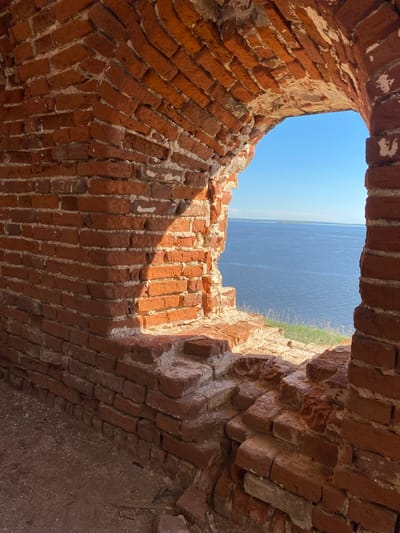 The image shows a view through a ruined brick structure towards water and the sky. The brickwork appears to be arched and forms a frame around the opening. Vegetation is visible on the horizon, above the water. The location is in Шуран, Russia.