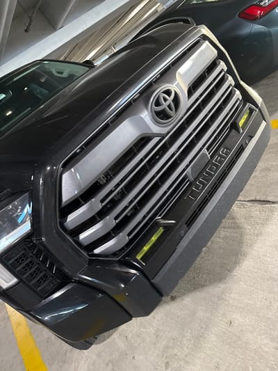 Captured in what appears to be a parking garage in the Hollywood Seminole Reservation, United States of America, the image focuses on the front grill of a black Toyota Tundra. The grill features the Toyota logo, horizontal silver slats, and "TUNDRA" lettering across the top. The vehicle is parked close to a concrete wall, marked with a yellow parking line. A portion of another vehicle is visible on the left side of the frame. The lighting conditions suggest an indoor environment.