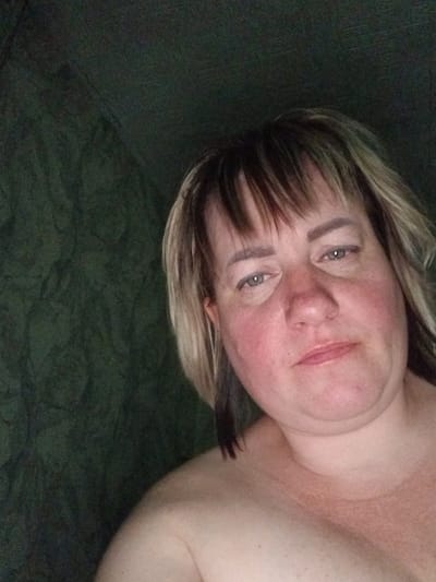 This is a close-up, front-facing selfie of a middle-aged Caucasian woman with blonde and brown hair with bangs. She has green eyes, fair skin with visible redness on her cheeks and chest, and is not wearing any visible clothing above the shoulders. The background is a dark green, textured surface, possibly fabric or a wall. The lighting is dim and appears to be coming from the front. There are no other people or objects clearly visible in the frame. The image does not contain any information about the location in Prokudskoye, Russia.