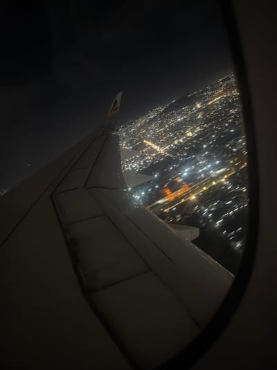 The image shows a view from an airplane window at night. The wing of the plane is visible, and through the window, the city lights of what is purported to be Addis Ababa, Ethiopia are seen. The city lights appear as scattered points of light, concentrated in patterns resembling streets and urban areas. The sky outside is dark.