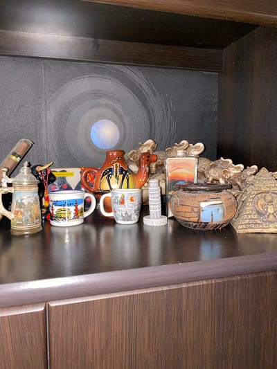 The image shows a drawer filled with various novelty items, likely souvenirs. The items are arranged vertically and include a miniature Eiffel Tower, a stylized Egyptian pyramid, and several mugs decorated with cityscapes and landmarks. One mug clearly depicts "BARCELONA," and another has Cyrillic text that reads "Петербург" (Saint Petersburg). There's also a decorative beer stein with an image of a European city. A small bull figurine is visible, along with what appears to be a cigar. The drawer itself has a dark, wood-grain finish on the right side, and the bottom of the drawer features a black surface with a circular, swirling pattern and a luminous element, possibly a light source or a decorative insert. The lighting suggests it is indoors, and the overall impression is that of a collection of mementos.