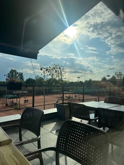 The image displays an outdoor patio overlooking multiple clay tennis courts in Giebułtów, Poland. The vantage point is from beneath a dark awning that extends from the upper left. Bright sunlight, accompanied by lens flare, emanates from the upper right quadrant of a blue sky scattered with white clouds.
In the immediate foreground, a patio area is furnished with several dark, modern-style chairs featuring perforated backs and square tables. A large dark pot containing a small, weeping tree with green foliage is centrally positioned on the patio. A smaller black planter with red flowers is attached to a railing on the left. The patio floor is composed of light-colored, possibly stone or concrete, tiles.
Beyond a clear railing, at least three red clay tennis courts are visible, each equipped with a net. Tall, dark floodlight poles are situated around the courts. Green mesh fencing encloses the courts, and multiple signs displaying "UTR PRO TENNIS" are affixed to this fencing. The background consists of dense green trees and foliage beyond the court perimeter. No individuals are present on the patio, courts, or within the discernible environment. The scene is static, depicting an unoccupied outdoor sports facility during daytime.