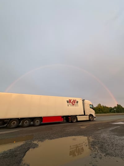 The image depicts a white semi-truck with a long white trailer parked on a gravel shoulder next to a paved road. A faint rainbow is visible in the overcast sky. The truck has the logo "KN" with the text "REFRIGERATED TRANSPORT" and a phone number "+49 1621 98 44-0" on the side of the trailer. The ground is wet, with puddles reflecting the truck and the surrounding area, suggesting recent rain. Trees and foliage line the distant side of the road. The lighting suggests it is either early morning or late evening, and the overall atmosphere is subdued due to the overcast weather. The scene appears to be a roadside rest stop or parking area, possibly in a rural or industrial zone, given the gravel and the truck. No people are visible in the image. The reflection in the water shows a mirror image of the truck, including the logo and text on the trailer.