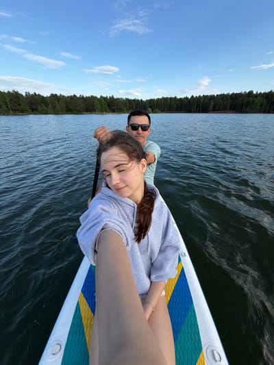 A man and a woman are lying on a paddleboard on a body of water. The man is wearing sunglasses and a light green shirt, and he is looking towards the camera. The woman is wearing a gray sweatshirt and has her eyes closed. The paddleboard is blue, yellow, and green. In the background, there is a line of trees and a blue sky with white clouds. The location is likely Selychka, Russia, given the request parameters. The couple seems to be relaxing and enjoying their time on the water.