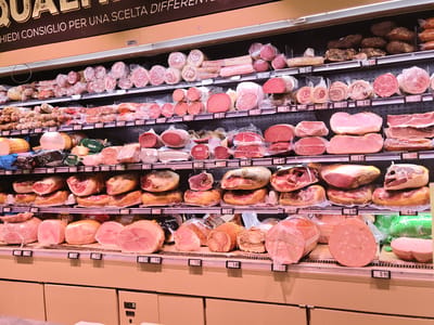The image shows a well-stocked display of cured meats in a supermarket. The cured meats are arranged on multiple shelves, with various types of salumi, hams, and other processed meats visible. Many of the products are packaged in clear plastic wrap, allowing their textures and colors to be seen. Some items are whole, while others are sliced or in larger portions. Price tags are visible in front of many of the products. The background shows more rows of similar products, suggesting a large deli section. The lighting is bright and consistent, typical of a retail environment. The overall impression is one of abundance and variety of meat products. The text above the display, visible in the top portion of the image, reads "QUALITA' CHIEDI CONSIGLIO PER UNA SCELTA DIFFERENTE," which translates to "QUALITY ASK FOR ADVICE FOR A DIFFERENT CHOICE." This suggests the location is a place where customers can seek guidance on their purchases. Given the text is in Italian and the context of a supermarket display, it is plausible this scene is from a supermarket in Quarto, Italy, or any other Italian city.