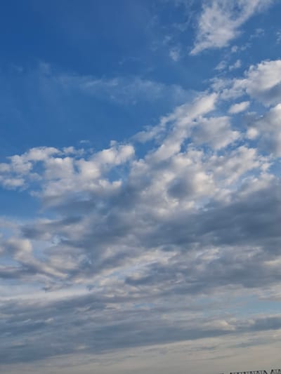 The image shows a cloudy sky with a blue hue. The clouds are varied in shape and density, with some appearing white and fluffy while others are gray and more solid. The lighting suggests it could be late afternoon or early morning. There are no visible people, subjects, or objects in the foreground or mid-ground of the image, only the sky and clouds. The bottom edge of the image shows a very faint hint of what appears to be a structure, possibly a bridge or some industrial construction, but it is too indistinct to identify. The image is a vertical, full-frame shot of the sky.