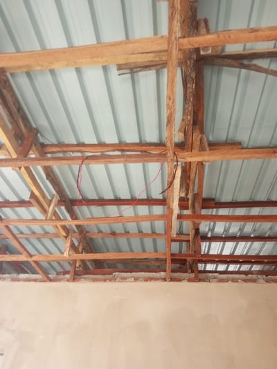 A cluttered interior shot reveals a wooden framework supporting a corrugated metal roof. Thin wooden beams are arranged in a grid pattern, intersecting with thicker, upright wooden supports. A red wire is haphazardly strung across the beams. Below the roof structure, a light-colored wall stretches across the frame's bottom, indicating the interior space. The image highlights the rustic and exposed construction of the building.