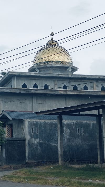 The image presents a mosque in Lonbaroh, Indonesia, under an overcast sky. The focal point is the mosque's large, golden dome, which is highly reflective and features intricate Arabic calligraphy in green script along its base. A crescent moon and star finial, a common Islamic symbol, tops the dome. Below the dome, white loudspeakers are mounted on the concrete structure, likely used for the call to prayer.
The mosque's main body is constructed from unpainted, rough grey concrete, featuring a series of recessed, arched windows. Utility cables or power lines traverse the upper portion of the frame, partially obscuring the dome and sky.
In the foreground and mid-ground, additional concrete structures are visible, including one with a corrugated metal roof supported by concrete pillars, forming a sheltered area. Another building section to the left has patterned tiles near an entrance. The ground level displays patches of green grass, dry earth, and some refuse, bordering a paved road or path. The overall setting suggests a local community environment. There are no people visible, and no specific activities are taking place. The subdued lighting from the overcast conditions implies either an early morning, late afternoon, or simply a cloudy day.
