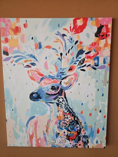 This is a medium close-up, eye-level, horizontal shot of a painting that appears to be a paint-by-numbers artwork of a deer's head. The deer's head is depicted in profile, facing right, with its body extending out of frame to the right and left. The artwork is characterized by a vibrant, abstract style with a palette of bright, pastel colors including pinks, blues, oranges, and yellows, interspersed with darker shades of navy and black. The deer's face is rendered with bold strokes of blues and pinks, and its eye is a dark circle. Its antlers and the area around its head are adorned with stylized floral and leaf motifs in a mix of colors. The background is a soft, pale blue with scattered dots and abstract shapes in various colors, suggesting an ethereal or dreamlike atmosphere. The painting is on canvas and is unframed, with its edges visible against a brown, textured surface, possibly a wall or table. There is no visible text in the image. The style is reminiscent of contemporary folk art or illustrative designs, with a focus on color and form rather than photorealism.