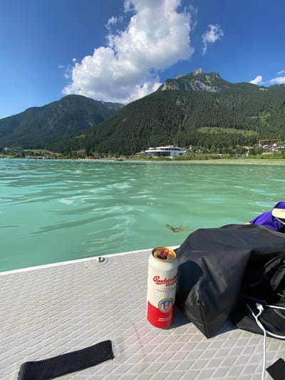 A Budweiser beer can rests on a paddleboard beside a black bag with a purple strap. The paddleboard is on a calm, turquoise body of water, likely a lake, given the mountain backdrop. Lush green forests cover the steep slopes of the mountains that rise dramatically from the water's edge. A small town or village with buildings is nestled along the shoreline, with a large, modern white building appearing to be a prominent structure. In the distance, the mountains continue to stretch, with some rocky outcrops visible on the higher elevations. The sky is a vibrant blue, dotted with fluffy white clouds. The sun appears to be high in the sky, suggesting it is midday. The scene evokes a sense of outdoor recreation and relaxation. The can of Budweiser has visible text, including "Budweiser," "Original Czech Lager," and "Owned by AB InBev Czech Republic." There are also red and blue crests on the can. The paddleboard has a textured, diamond-patterned surface and a metal D-ring and a black handle or strap.