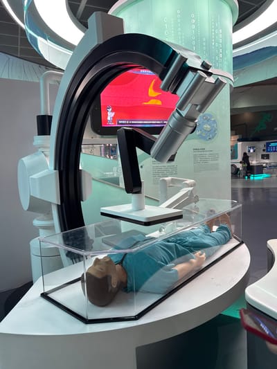 An indoor exhibition hall in Guangming District, China, presents a multi-component display focused on medical technology. Centrally, a life-size male mannequin, dressed in teal scrubs, lies supine on a white curved platform, contained within a transparent acrylic case. Above the mannequin, a robotic arm-like structure, featuring a flat, dark rectangular sensor, extends downward from a large, stylized white and black arched apparatus, resembling an advanced medical imaging device. In the immediate background, a large curved display screen exhibits animated graphics on a red background, depicting internal biological imagery alongside a small cartoon figure and Chinese text. To the right, a tall, illuminated column features falling white binary code (0s and 1s) graphics on a green backdrop. Adjacent to this, an informational panel displays Chinese text and diagrams referencing "Drug Design and Development." The floor is light-colored and reflective. In the blurred distant background, indistinct human figures, including one appearing to push a stroller, are visible. A portion of a red mobile phone case is visible in the bottom right foreground.