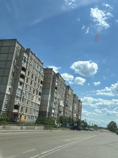 The image captures a street scene in Balakliya, Ukraine, on a bright, sunny day. The dominant subjects are a row of multi-story, concrete apartment buildings on the left, an asphalt road on the right, and a vivid blue sky dotted with white cumulus clouds overhead.
The apartment buildings exhibit typical Soviet-era architecture, characterized by their grey concrete facades and numerous square windows, some with small balconies or air conditioning units. Many windows are open, and several satellite dishes are mounted on the exterior. The buildings appear worn, suggesting age, though a section of one building has been painted yellow, possibly indicating a ground-floor commercial establishment. The number "123" is clearly visible on this yellow section.
The sky is a deep blue, filled with scattered white clouds, some appearing fluffy and others more wispy. The bright sunlight casts no significant shadows, indicating midday or early afternoon. A faint reddish smudge is visible in the upper left portion of the sky, likely a lens artifact.
To the right of the buildings, a paved, two-lane road runs parallel. It features double solid white lines down the center, implying traffic in both directions or a no-passing zone. Several cars, mostly dark-colored sedans and SUVs, are parked along the right side of the road. Further down the street, more vehicles and distant trees are visible, suggesting a continuation of the urban environment. A streetlight pole stands near the parked cars, and a wire fence separates the buildings from the road in parts. A small section of a blue and yellow flag is faintly visible near the buildings on the left side, confirming the Ukrainian context. A partial sign on the right side, near the parked cars, shows the letters "БЕТО..." and some unreadable text below it.
