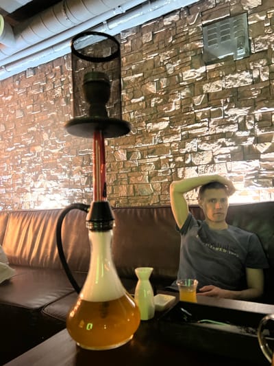 The scene appears to be an indoor lounge in Zaosinovo, Russia. A young person reclines on a dark leather sofa adjacent to a coffee table with a glass of liquid. A hookah is set up next to the sofa. The background features a wall decorated with stone-like tiles. In the top left of the image is a ventilation unit on the wall. The sofa is the point of reference in the bottom right corner.