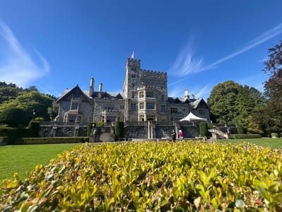 The image shows a large, stone castle-like building under a bright blue sky with wispy white clouds. The building has a tall central tower with battlements and a Canadian flag at its peak. It features multiple gabled roofs, chimneys, and large windows. In front of the building is a manicured lawn and hedges, with a low-lying hedge in the foreground that is out of focus. There are some people standing on a raised walkway in front of the building. The castle is situated on a gentle slope, with mature trees visible on either side and in the background. This appears to be the Royal BC Museum in Langford, Canada.