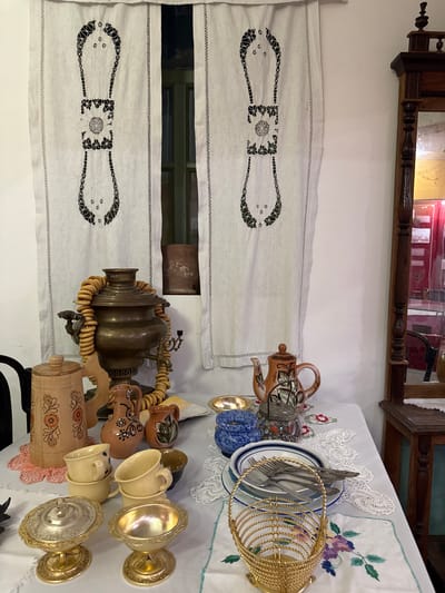 This image depicts a still life of a table set for a traditional tea ceremony, likely in a museum setting in Fort-Shevchenko, Kazakhstan. The foreground features a white tablecloth adorned with embroidered flowers. On the table are various objects, including a large brass samovar draped with a string of dried bread rings, several ceramic cups and pitchers, a wooden cylindrical container with painted floral designs, a decorative ceramic teapot, a glass jar filled with small objects, and stacks of plates with silverware. Two ornate gold-colored bowls or chalices are prominently displayed. In the background, white curtains with black embroidered patterns hang over a window. To the right, a dark wooden antique cabinet with a mirror is partially visible, reflecting a portion of the room. There are no people present in the image.