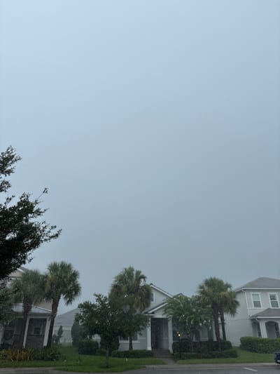 This media file depicts a residential street scene in Bradenton, Florida, during a rain shower. The perspective is from a low angle, looking upwards towards a hazy, overcast sky. The houses visible are of modern architecture, with neutral-toned exteriors and pitched roofs. One house features a light gray facade with white trim and multiple windows. Another house, partially visible, has a white exterior with dark shutters and a covered porch.
Lush green landscaping, including manicured hedges and palm trees, surrounds the properties. The palm trees, a characteristic element of the Florida landscape, are swaying in the wind and appear somewhat windswept due to the weather. Small white dots, likely raindrops, are visible throughout the image, indicating that it is actively raining. The sky is a uniform, pale gray, suggesting either an early morning, late afternoon, or a heavily overcast day. There are no people or vehicles prominently featured in the image. The overall impression is one of a quiet, somewhat gloomy day due to the rain. No visible text is present in the image.