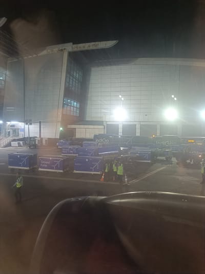 This is a photograph taken at night from an airplane window, looking out onto an airport tarmac in Agassaim, India. The foreground is dominated by the dark, curved surface of an airplane engine nacelle.
Beyond the nacelle, the tarmac is illuminated by bright overhead lights, casting a harsh glow on the scene. A line of large, blue cargo containers, branded with "IndiGo" and "Ground Force," are visible. Some of these containers are on wheeled dollies.
Several individuals, dressed in high-visibility vests in bright yellow and green, are present on the tarmac, seemingly engaged in ground operations related to the cargo. One person in a yellow vest is walking away from the camera towards the left. Others are clustered near the cargo containers.
In the background, a modern airport terminal building is visible. It features a large, white, tiled facade and expansive windows, some of which are illuminated from within. The overall impression is of a busy, operational airport at night, with cargo being loaded or unloaded.