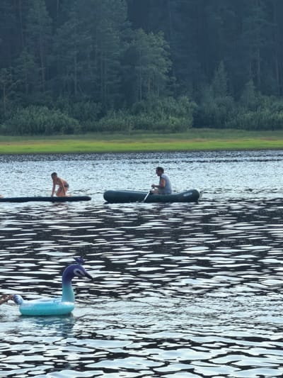 In the city of Sysert, Russia, on a body of water, a man is paddling an inflatable boat with a single oar. In the background, a person kneels on an inflatable raft. Closer to the foreground, an inflatable toy shaped like a peacock floats on the water. The scene is framed by a dense forest on the distant shore.