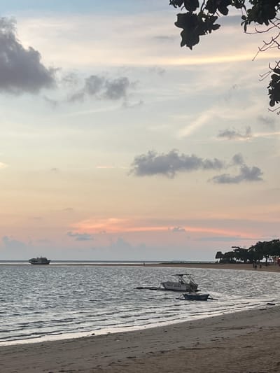 The image depicts a serene beach scene at dusk in Cuyo, Philippines. The sky is painted with soft hues of pink, orange, and pale blue, with scattered clouds. In the distance, a large cargo ship is visible on the horizon. Closer to the shore, two smaller boats are anchored in the calm water, one with "LIFE GUARD" written on its side. The beach is sandy, with the gentle waves of the sea lapping at the shore. The silhouette of trees and a few people walking can be seen along the coastline in the background, adding a sense of quiet activity to the peaceful atmosphere.