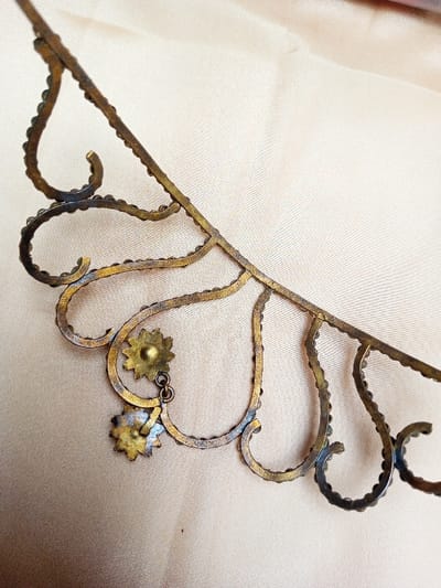 A close-up photograph captures an aged, ornate metal object, possibly a piece of jewelry or decorative trim, displayed on a light beige fabric surface in Qarabucaq qəsəbə, Azerbaijan. The object, appearing to be crafted from an oxidized brass or bronze alloy, exhibits a curvilinear, openwork design. Its main structure comprises a series of interconnected, stylized heart-shaped or scalloped elements along a horizontal bar, each outline adorned with numerous small, dark, bead-like protrusions. Two small, seven-petaled, flower-shaped charms are suspended via small rings from one of the curved elements on the left side of the frame. The metal shows significant tarnish, varied patination from dark brown to dull gold, and hints of green oxidation in recessed areas. The background is an indistinct, lightly textured, cream-colored fabric, possibly satin or silk, with subtle folds. No people or specific actions are visible.
