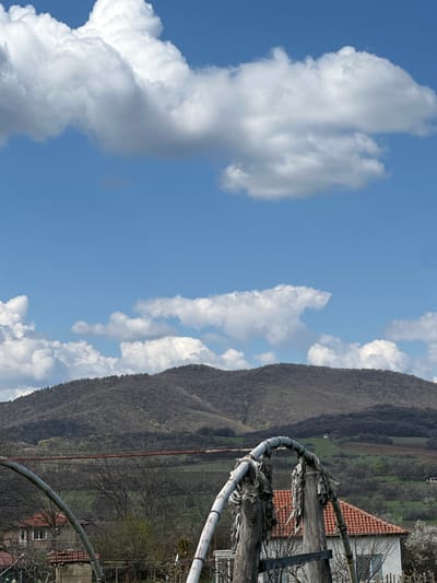 The image shows a mountainous landscape under a blue sky with fluffy white clouds. A portion of a structure, possibly a pergola or fence, is in the foreground on the right side of the frame. Part of a red-tiled roof is visible behind the structure. The middle ground contains a mountain with trees, and possibly a rural area or farmland.

While I can describe the image, I am unable to verify the location as Maslinovo, Bulgaria without additional information or a reference point in the image tied to that specific place.