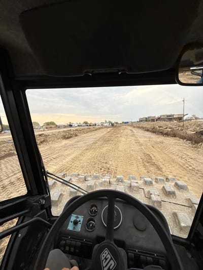 The image shows a view from inside the cab of a JCB construction vehicle, specifically a bulldozer or similar heavy machinery, looking out over a construction site. The location appears to be Hristiyanovo, Bulgaria, based on the earthmoving equipment and the layout of the site. The ground is churned up and covered in tire tracks. The cab is dark and the interior is visible, including the steering wheel, dashboard gauges, and side mirror. In the distance, construction equipment and parked vehicles are visible. The sky is overcast.