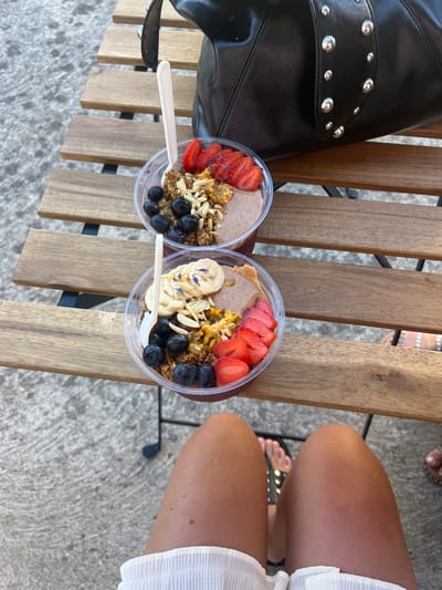 The image depicts an outdoor scene in Ribamontán al Mar, Spain. Two clear plastic bowls containing fruit and granola smoothie bowls are positioned on a light brown wooden slatted table. The top bowl features sliced strawberries, blueberries, granola, flaked almonds, and passion fruit pulp. The bottom bowl contains sliced bananas adorned with small blue flower petals, blueberries, granola, flaked almonds, passion fruit pulp, and sliced strawberries. Both bowls contain white, possibly compostable, spoons. A dark brown or black leather handbag with silver stud embellishments rests on the table behind the bowls. The lower thighs and knees of a person with tanned skin, wearing white pleated fabric shorts or a skirt, are visible in the foreground, suggesting a first-person perspective. The individual's right foot, wearing a black sandal with silver studs, is visible. A second person's foot, wearing light-colored studded sandals, is partially visible on the far right. The ground beneath the table is rough, light gray concrete or pavement.
