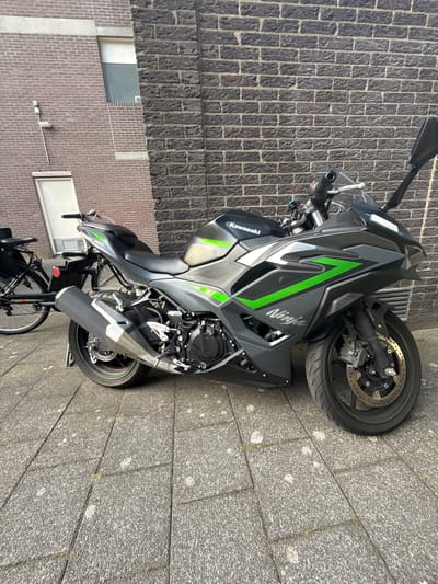 A Kawasaki Ninja motorcycle is parked on a grey stone sidewalk. The motorcycle is dark grey with lime green accents and decals. It is a sportbike with a sleek design. The motorcycle is positioned diagonally to the right of the frame. A bicycle with a black rear rack is parked behind the motorcycle. The background consists of a dark brick wall and a lighter brick wall with a white door and a window. A light fixture is mounted on the lighter brick wall. The scene appears to be in an urban setting. The context of Amstelveen, Netherlands is provided, but no specific landmarks or identifying features within the image confirm this location.