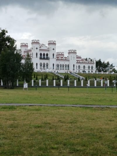A wide, low-angle shot captures a grand, white, multi-turreted castle structure situated on a grassy hill under a cloudy sky. The castle exhibits neo-Gothic architectural elements, with numerous pointed arch windows and decorative crenellations on its towers. A white, ornate fence with evenly spaced pillars runs along the base of the hill, separating the castle grounds from the foreground. A manicured lawn with decorative shrubbery and lampposts is visible behind the fence. Several individuals are scattered on the stairs leading up to the castle and walking on the grounds, suggesting the location is open to visitors. In the foreground, a well-maintained grassy field extends towards the viewer, with a paved path bordering the lower edge of the frame. The overall impression is that of a historic, well-preserved landmark in a park-like setting. The castle is the Koźlakow Palace, located in the village of Ivyanets, within the Valozhyn District of the Minsk Region, Belarus. The coordinates for this location are 53°53'28.8"N 26°43'25.2"E.