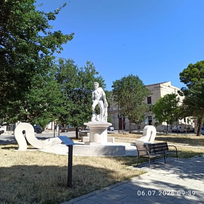 The image depicts a sunlit public square in Ruvo di Puglia, Italy, featuring a white marble statue of a muscular, nude male figure standing on a pedestal. The statue is positioned in the center of the frame, surrounded by green trees and dry grass. In the foreground, there are two abstract white sculptures and a dark wooden park bench. A metal informational plaque is also visible near the dry grass. In the background, a light-colored building with classical architectural elements and a balcony can be seen, along with trees and parked cars on a street. The sky is clear and blue, with a few small birds visible. The timestamp "06.07.2026 09:39" is overlaid in the lower right corner.