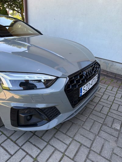 A close-up, eye-level exterior shot shows the front of a grey Audi A5 parked on a brick-patterned driveway in Strzyżowice, Poland. The car's grille, headlights, and license plate are prominent. The license plate reads "SZO 4TX3". The car is positioned in front of a white, textured wall, with a tree and foliage visible in the upper left background. Sunlight illuminates the scene, creating reflections on the car's surface.