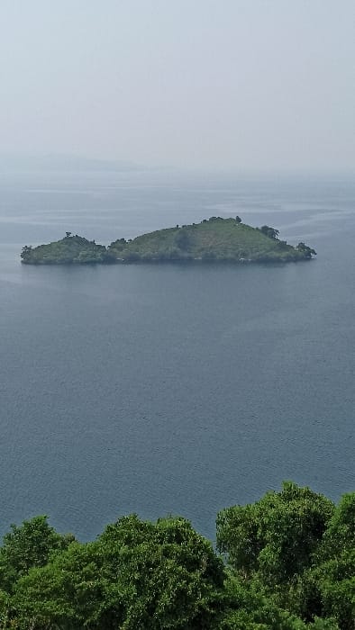 The image shows an aerial view of a body of water with a long, narrow island. The island is covered in green vegetation. The water is calm and reflects the sky. The right side of the image features a thick cluster of green trees. The scene is generally peaceful and serene.