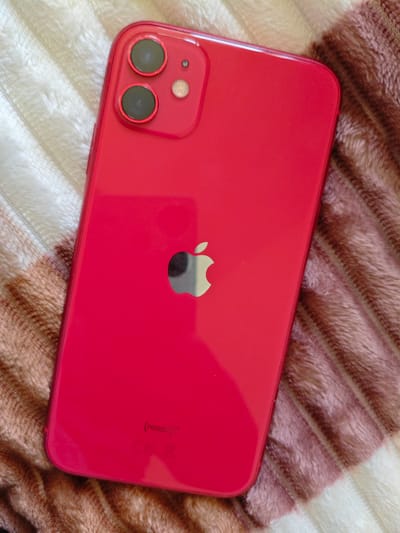 The image shows the back of a red iPhone 11. The phone is lying on a textured surface that appears to be a blanket or comforter with stripes of white and brown. The Apple logo is visible in the center of the phone. The camera module, which consists of two lenses and a flash, is located in the upper left corner of the phone. There are also some markings on the bottom of the phone, including "PRODUCT (RED)" and regulatory symbols. There is no indication of the city of Sumgayit, Azerbaijan in the image.