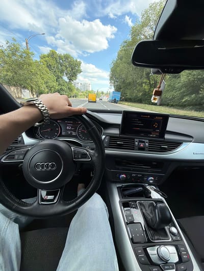 This media file shows a first-person view from the driver's seat of an Audi A6, likely in Orhei, Moldova, on a sunny day with scattered clouds. The driver's hands are visible on the steering wheel, and they are wearing a silver watch. The car's dashboard displays the current speed as 62 km/h and the time as 16:02. The infotainment screen shows "Источник" (Source) and "Настройки" (Settings), with a music icon. Outside the car, trees and buildings are visible, along with a yellow van on the road ahead. The scene suggests the person is actively driving during the afternoon. The "quattro" badge on the dashboard and "S line" emblem on the steering wheel indicate the car's model and trim. A car air freshener is hanging from the rearview mirror.