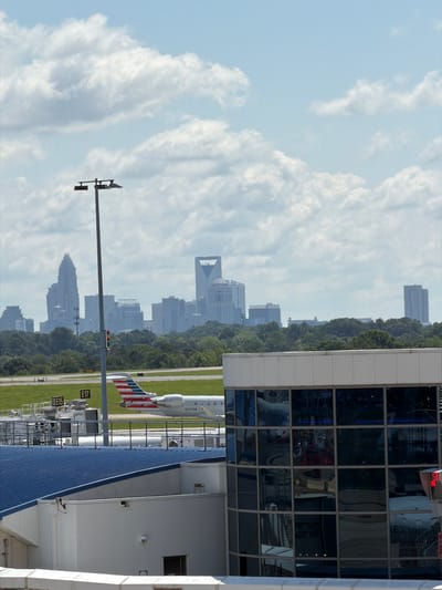 The image depicts a cloudy sky over an airport. In the foreground, a modern building with large glass windows reflects the sky and a cityscape. The building's exterior is predominantly white with a blue, corrugated roof section visible on the right. Several panes of glass in the building are tinted, offering glimpses of what appears to be an interior space.

To the left of the building, a tall lamppost stands, a common fixture at airports. In the middle ground, an airplane with the American Airlines livery is parked on the tarmac. The airplane's tail fin, adorned with red, white, and blue stripes, is clearly visible. The airport runway stretches out behind the plane, with grass on either side.

In the background, a sprawling cityscape with numerous tall buildings rises above a line of lush green trees. The architecture of the city suggests a significant urban center. The sky is filled with large, billowing white clouds, indicating partly cloudy weather. The lighting suggests it is daytime, though the exact time is not discernible. The overall scene conveys the bustling atmosphere of an airport juxtaposed with a distant urban skyline, set against a backdrop of a dynamic sky. The image is oriented vertically, with the cityscape and airport features appearing sideways. Visible text on the lamppost appears to be "E17" and "E15".