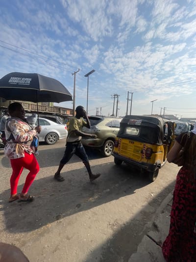 The image depicts a street scene in Akowonjo, Nigeria, characterized by a bright sky with scattered clouds. In the foreground, a woman wearing red leggings and a patterned top walks while holding a black umbrella open. She appears to be carrying a baby or a child on her back, though it is partially obscured. To her right, a man in a green t-shirt and dark shorts walks briskly, looking to his right and touching his face. Behind them, a yellow auto-rickshaw, identified by "TVS" on its rear, is parked or moving slowly. Several other vehicles, including a silver SUV and other cars, are visible in the background, along with utility poles and buildings. The ground is paved with concrete and appears somewhat worn. The overall impression is of a busy urban environment during daylight hours.