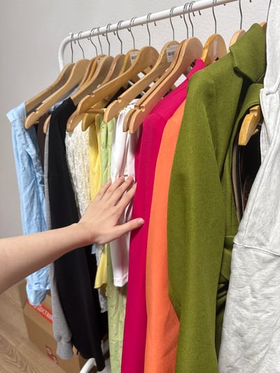 In Drozhzhino, Russia, a rack of clothing is displayed. The rack holds numerous items of clothing in various colors, including blue, black, white, yellow, green, orange, and pink. A person's hand is visible, touching one of the garments. In the background, a cardboard box with Russian text and a part of a wall are visible. The scene appears to be indoors, likely in a residential or retail setting.