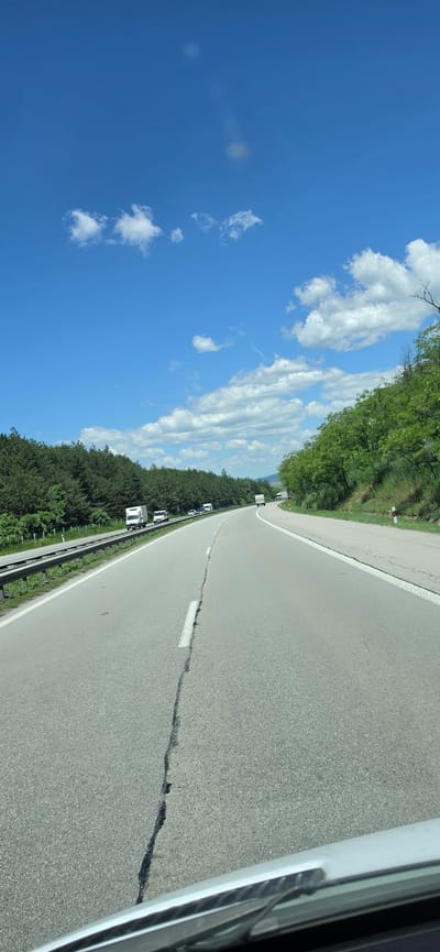A roadside scene, likely Gorna Vasilitsa, Bulgaria. The perspective is from inside a vehicle moving along a cracked asphalt road with a white line divider. The road is flanked by dense green trees on one side and a guardrail. Several vehicles, possibly trucks, are visible further down the road. The sky is bright blue with scattered cumulus clouds.