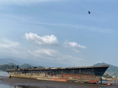 A wide shot depicts a large, weathered wooden boat or barge beached on a muddy or sandy shore under a bright, partly cloudy sky. The vessel features a long, dark hull with a distinct red-painted waterline along its lower section, indicating its extensive use. Its bow points towards the right of the frame, resting firmly on the ground, while the rest of the boat sits partially in shallow water or mud.
Several people are visible around the boat. On the left, a person is seated on the ground near the boat's hull, appearing to be working or observing. Further along the left side, another figure stands on the boat's deck or edge. A third person is faintly visible near the boat's bow on the right, possibly attending to ropes or supports.
The background is dominated by lush green mountains rising dramatically against the horizon, characteristic of coastal landscapes like Victoria, Cameroon. The sky above is clear blue with scattered white cumulus clouds. Two dark birds are in flight high overhead, adding a sense of scale. The foreground consists of wet, dark ground, possibly sand or mud, with reflections of the sky in shallow water. The scene suggests a busy but tranquil coastal or riverside environment, likely during daylight hours with fair weather. No visible text is present in the image.