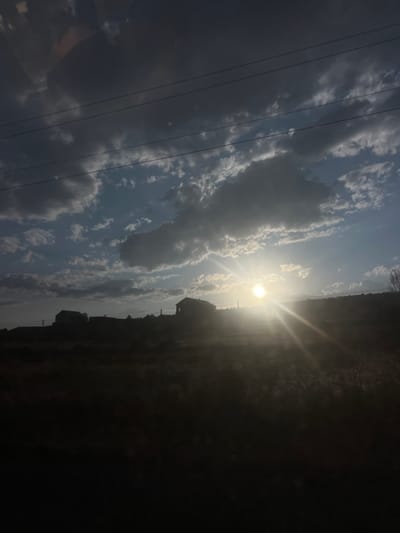 A landscape image features a bright sun shining through a cloudy sky. Power lines run diagonally across the left side. The right side is dominated by dark shadows, obscuring details of the terrain and vegetation. Silhouetted buildings are visible along the horizon line where the sun is positioned. The overall scene is a high-contrast view of a landscape bathed in sunlight.