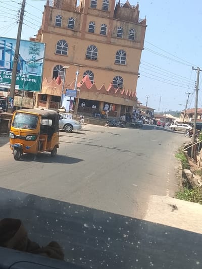 The image captures a daytime street scene in Iragbiji, Nigeria. A prominent five-story building with a tan-orange facade, featuring multiple arched windows and a decorative crenellated roofline, is positioned on the right side of a two-lane asphalt road. On the ground floor of this building, an open porch area hosts approximately five individuals who are either standing or seated, with additional figures visible further inside.

In the left foreground, a yellow and black motorized tricycle (keke napep) is in motion on the road, traveling away from the viewer. Behind it, a silver sedan is parked on the roadside. Multiple motorcycles are parked along the curb in front of the large building. The road continues into the background, where additional vehicles, including cars and motorcycles, are observed.

Utility poles with a dense network of overhead wires run along both sides of the street. On the left, a large blue and green billboard is visible, partially obscured, with text indicating "M. OF AGRICULTUIRE, IRAGBIJI (FJADSI)." A concrete drainage ditch lines the right side of the road, bordered by sparse vegetation. The sky is clear blue, and the lighting suggests midday, evidenced by relatively short shadows. The foreground and lower portion of the image contain numerous small white specks, consistent with dust or debris on a transparent viewing surface from which the photograph was taken.