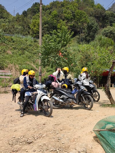 In Phố Bảng Commune, Vietnam, four people in yellow helmets are standing near three parked motorbikes on a dirt road. The people appear to be taking a break, as one is adjusting her helmet. Behind them, a lush green hillside rises. To the right is a tree and a glimpse of a red awning. In the foreground, a green net lies on the ground. A sign post partially visible on the left. Overhead power lines cross the upper portion of the image.