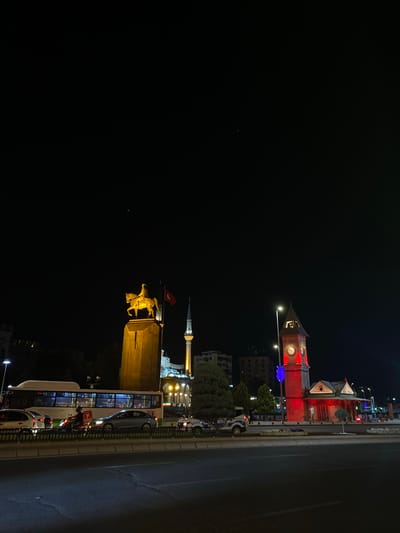 This is a nighttime outdoor shot of the Kayseri Clock Tower and the Atatürk Monument in Kayseri, Turkey. The monument, a golden equestrian statue of Mustafa Kemal Atatürk, stands prominently on a raised pedestal, illuminated by a warm light. Beside it, a minaret of a mosque rises into the dark sky. To the right of the monument is the distinct red-illuminated clock tower. In the foreground, a busy street with traffic is visible, featuring cars, a bus, and a motorcycle with a rider. A police car is parked on the side of the road. The scene is set against a dark night sky with some distant city lights.